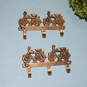 May include: Two sets of bronze-colored metal wall hooks. Each set features a decorative design of two bicycles side-by-side. Each hook set has three hooks for hanging items. The hooks are mounted on a light blue surface.