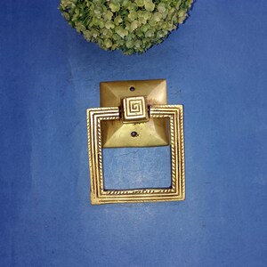 May include: A gold square door knocker with a decorative geometric design. The knocker is mounted on a blue surface.