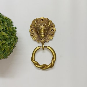 Peut inclure: Un porte-serviettes mural de couleur laiton. L'anneau est fixé à une tête de taureau décorative avec un motif floral. La tête de taureau est centrée sur un fond blanc. L'anneau à serviette a un design de corde torsadée.