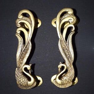 Könnte beinhalten: Zwei verzierte Türgriffe aus Messing. Jeder Griff zeigt einen Schwan an der Basis und ein stilisiertes Federdesign, das sich nach oben wölbt. Die Griffe sind goldfarben und auf dunklem Hintergrund montiert.