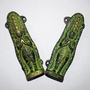 May include: Two decorative, green and gold metal handles with a detailed relief design. The handles feature a central figure with ornate detailing and a dark green background. Each handle has a mounting bracket.