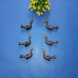 Op de afbeelding: Zes bronzen pauwvormige trekhandgrepen gerangschikt op een blauw oppervlak. Elke trekker heeft een gedetailleerd pauwontwerp met een decoratieve kuif. Een kleine opstelling van gele en groene kunstbloemen bevindt zich in de bovenhoek.