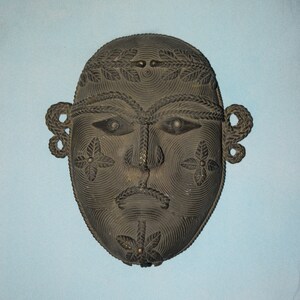 Può includere: Una maschera in legno nero con intagli intricati di un viso, foglie e motivi geometrici. La maschera ha una forma arrotondata ed è decorata con piccoli accenti metallici.