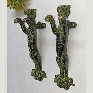 Pode incluir: Duas maçanetas de porta em estilo antigo em forma de tigre. As maçanetas são pretas com detalhes verdes e dourados, montadas em uma superfície branca. As patas do tigre estão estendidas e as maçanetas são muito detalhadas.