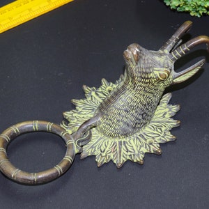 May include: A brass door knocker featuring a detailed design of an animal head with antlers. The knocker has a ring for pulling and is mounted on a decorative base.