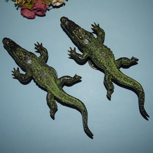 May include: Two green plastic alligator figurines with black eyes and gold accents. The alligators are posed with their mouths closed and their tails curled.
