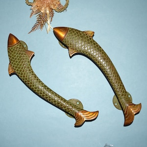 May include: Two decorative door handles in the shape of fish. The handles are olive green with gold accents on the head, fins, and tail. The fish scales are detailed. The handles are mounted on a light blue surface.