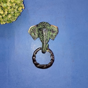 May include: A brass door knocker shaped like an elephant's head with a green patina. The knocker has a twisted ring for pulling.