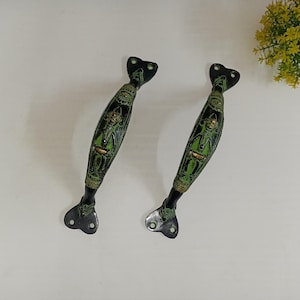 Könnte beinhalten: Zwei schwarze und grüne Metall-Türgriffe mit einem dekorativen Design einer Person mit Hut. Die Griffe haben eine herzförmige Basis.
