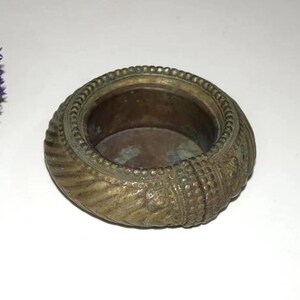 May include: An antique brass bowl with intricate embossed patterns around the rim. The bowl has a circular shape with a wide opening and a slightly textured surface. The color is a mix of gold and brown, suggesting age and use.