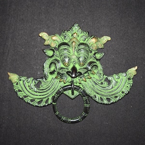 May include: A green and gold metal door knocker with a detailed design of a mythical creature's face. The knocker has a ring for pulling.