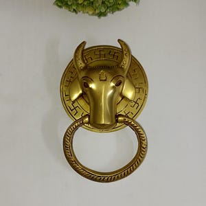 Puede incluir: Un llamador de puerta de latón de color dorado con forma de cabeza de toro con un anillo circular. La cabeza de toro tiene un símbolo de esvástica en la frente y un anillo alrededor del cuello.