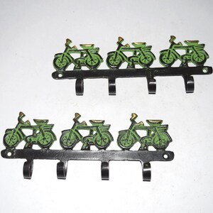 May include: Two sets of decorative wall hooks. Each set features a row of three black hooks with green bicycle silhouettes and gold accents. The hooks are mounted on a black metal base.