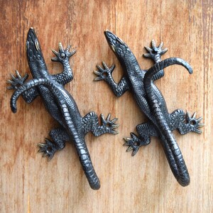 May include: Two dark gray metal lizard-shaped door pulls or cabinet handles. The lizards are detailed with textured scales and outstretched feet. The handles are mounted on a light brown wooden surface.