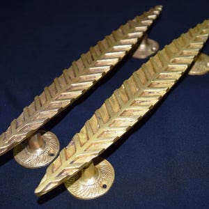 Könnte beinhalten: Zwei Türgriffe aus Messing mit Blattmuster. Die goldfarbenen Griffe haben eine strukturierte Oberfläche. Jeder Griff ist etwa 30 cm lang und auf einer kreisförmigen Basis montiert.