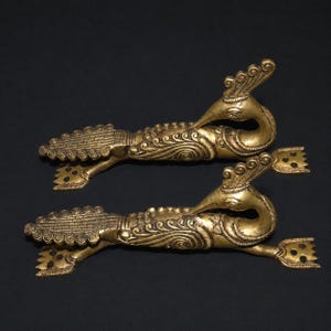 Puede incluir: Dos objetos metálicos dorados ornamentados con forma de pavo real. Cada uno tiene un cuerpo detallado con patrones arremolinados, un cuello largo y una cabeza con corona. Los objetos tienen un extremo decorativo en forma de abanico, sobre fondo negro.