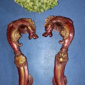May include: Two ornate, red and gold door handles with a dragon-like design. The handles are curved and feature intricate detailing, including a dragon head at the top and decorative elements along the length. The handles are set against a blue background.