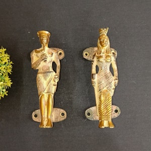 May include: Two gold-toned door handles with decorative figures. The handle on the left features a male figure with a crown and a staff. The handle on the right features a female figure with a crown and a staff.