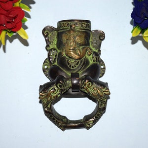 May include: Green and gold metal door knocker featuring a detailed sculpture of the Hindu deity Ganesha. The knocker is shaped like a ring with a Ganesha figure sitting on top.