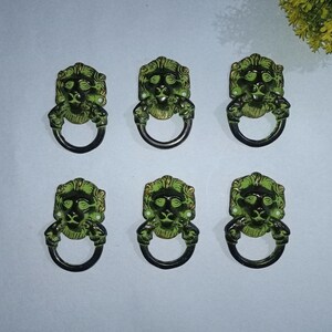 May include: A set of six metal drawer pulls, each featuring a lion head design with a circular ring handle. The hardware has a dark green and black finish with metallic gold highlights on the lion's mane and facial features.