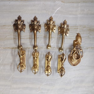 May include: A collection of five brass hardware pieces including four door handles featuring an elephant head design paired with vajra-shaped pulls, and one circular door knocker featuring a bird figure perched atop a decorative base.