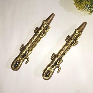 May include: Two antique brass-coloured alligator-shaped drawer pulls. Each pull features a detailed design with a textured body, small legs, and a curved tail. The pulls are positioned on a white surface.