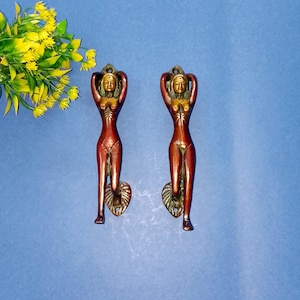 Puede incluir: Dos tiradores de puerta en tono bronce en forma de figuras femeninas estilizadas con los brazos levantados. Las figuras tienen un acabado marrón rojizo y están montadas sobre un fondo azul. Un ramo de flores amarillas está en la esquina superior izquierda.