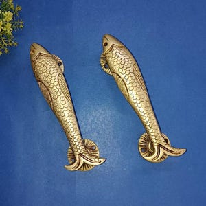 May include: Two brass-coloured fish-shaped door handles. The handles feature detailed scales and fins, set against a blue background. These decorative handles are suitable for a nautical or coastal theme, and are functional.