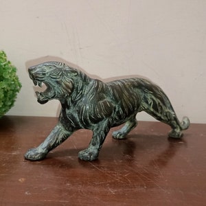 May include: A green bronze figurine of a snarling tiger. The tiger is standing on all fours with its mouth open and its teeth bared. The figurine is detailed with realistic features and a textured surface.