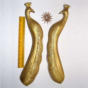 May include: Two gold-colored peacock-shaped decorative handles with intricate embossed detailing. A yellow ruler is placed next to one handle. A star-shaped ornament with a central gem is between the handles. The handles are approximately 10 inches long.