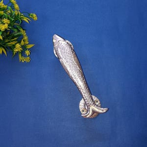 May include: A silver fish-shaped door handle with detailed scales and fins, mounted on a round base. The handle is set against a blue background. The handle is a decorative item.