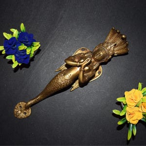May include: A detailed bronze mermaid door handle, featuring a scaled tail and a crown. The handle is accompanied by blue and yellow floral arrangements against a dark backdrop. The mermaid is holding a small creature.