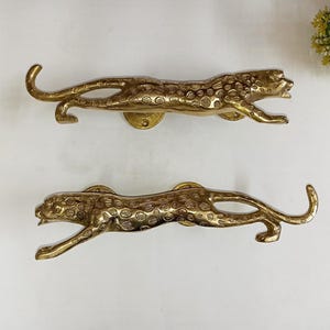 May include: Two brass-colored leopard-shaped door pulls. The door pulls are in a running pose, with detailed spots and a circular mounting plate. The hardware is made of metal and is suitable for furniture or cabinet doors.