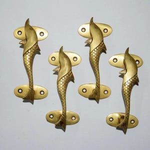May include: Four brass door handles, each shaped like a stylized fish. The handles have a fish-shaped grip and are mounted on oval plates with screw holes. The fish have detailed scales and fins.