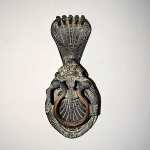 May include: Antique bronze door knocker featuring a cobra design. The knocker has a detailed cobra hood at the top, a decorative face, and a copper-colored ring for knocking. The overall design is ornate and detailed.