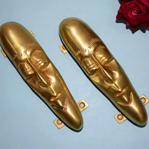 May include: A pair of gold-coloured, elongated face sculptures with detailed facial features. Each sculpture has a nose, eyes, and mouth. The sculptures are mounted on a light blue surface, with a red rose in the background.