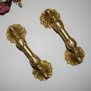 May include: Two antique brass door handles. Each handle features a central bar with floral accents and is attached to a decorative, flower-shaped base. The handles are a warm gold color and appear to be made of metal.
