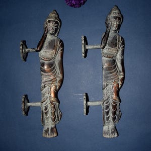 May include: Two bronze-effect Buddha figure door pulls. Each figure is in a standing pose with one hand raised to the head. The pulls have a decorative circular handle. The background is a dark blue. Purple flowers are in the upper right corner.