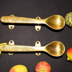 May include: Two gold-colored spoons with intricate designs on the handles are mounted on a black surface. The spoons are positioned horizontally, with the bowls facing upwards. There are three fruits, a yellow pear, a red apple, and a green melon, placed on the surface below the spoons.