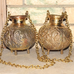 May include: Two ornate brass hanging pots with intricate floral designs and three legs. Each pot has a chain attached to the top.
