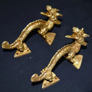 以下が含まれることがあります： 2つの豪華な金色のドラゴンドアハンドル。各ハンドルは、湾曲した体とネジ穴のある三角形のベースを備えた、詳細なドラゴンの姿を特徴としています。ハンドルは暗い背景に配置されています。