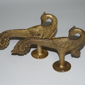 May include: Two brass peacock-shaped decorative hooks. The hooks are detailed with engraved feathers and stand on round bases. The peacocks have curved necks and are facing opposite directions. Ideal for adding an elegant touch to any space.