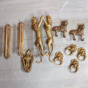 May include: A collection of brass hardware including two rectangular bar pulls, two leopard-shaped handles, two cat-shaped double hooks, one eagle-shaped door knocker, and three lion-head ring pulls, all featuring a polished metallic gold finish.