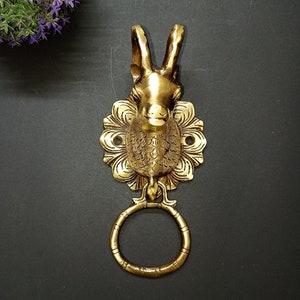 May include: A gold-toned door knocker featuring an intricate design of a goat's head with horns and a floral pattern. The knocker has a circular ring for knocking.