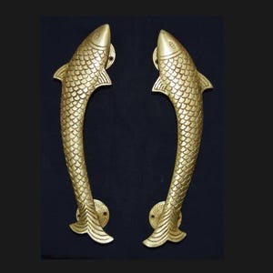 May include: A pair of gold-coloured brass door handles, each crafted in the shape of a fish. The handles feature detailed scale patterns and fin designs. They are mounted on decorative circular plates.