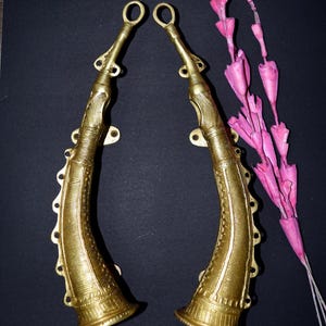 May include: Two decorative, gold-coloured, horn-shaped objects with detailed patterns. They are arranged symmetrically against a dark background, with a cluster of pink, paper-like flowers to the right.