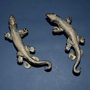 Op de afbeelding: Twee antieke bronzen deurgrepen in de vorm van een hagedis met een gestructureerd oppervlak. De hagedissen bevinden zich in een gebogen positie met hun staarten om hun lichamen gewikkeld.