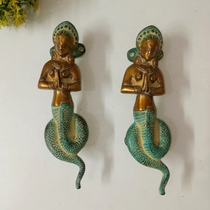 May include: A pair of ornate brass door handles featuring a woman figure with a snake tail. The woman is depicted with her hands clasped in prayer and is adorned with jewelry. The snake tail is curled around her legs and has a green patina.