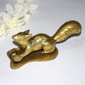 May include: A golden squirrel figurine in a relaxed pose, resting on a leaf-shaped base. The sculpture has detailed fur texture and a curved tail. The squirrel is a warm, metallic gold color, and the overall design is elegant.