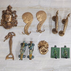 Op de afbeelding: Een collectie messing beslag, waaronder een decoratieve wandplaat met een gezicht, twee wandhaken met vrouwenprofielen, twee pauwvormige handgrepen, een zeemeermin-handgreep, twee Egyptische figuren, een ronde deurklopper en twee groene vierkante haken.